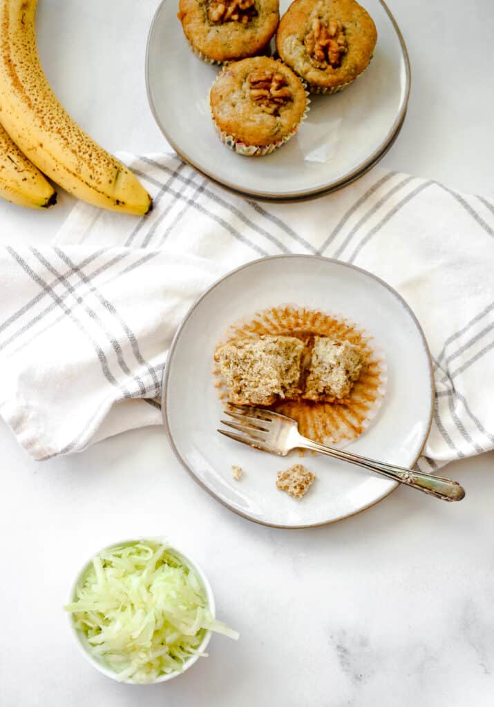 Banana zucchini muffin on a small plate with shredded zucchini and extra muffins on the side.